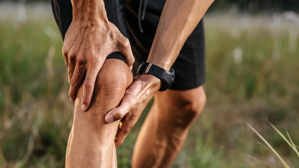 Deportista sujetando su rodilla con ambas manos por una lesión o calambre durante el entrenamiento al aire libre.
