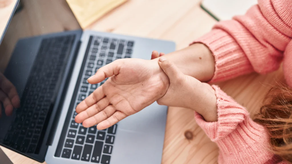 Mujer joven con suéter rosa sujetando su muñeca con dolor frente a una computadora portátil.